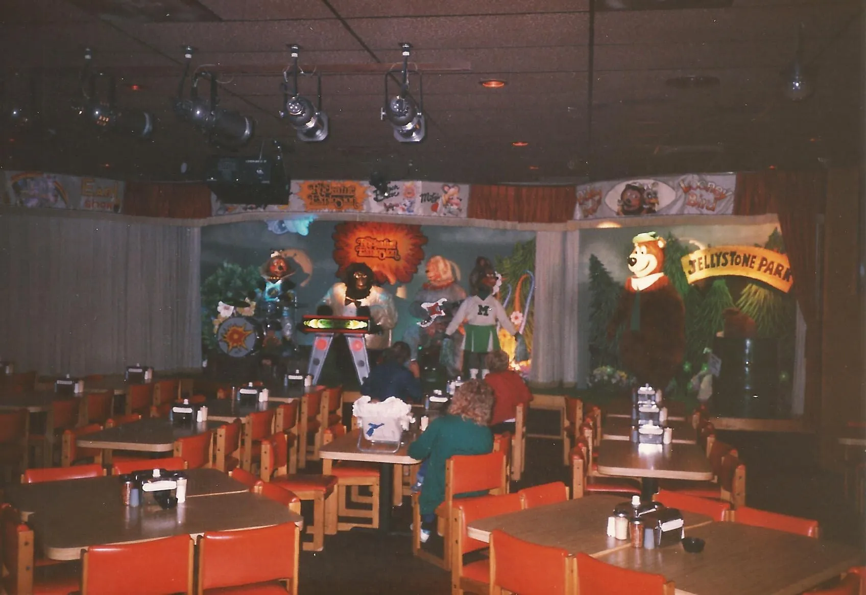 A ShowBiz Pizza Place showroom with the animatronic band on stage, the Jellystone Park stage at right, and families seated at the tables.